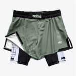 A pair of sage green two-in-one athletic shorts with a black and white inner compression layer, featuring grid patterns and stylized branding.