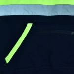 Close-up of a navy blue safety hoodie featuring a kangaroo pocket with neon yellow trim, a horizontal zipper pocket, a reflective silver stripe, and a neon yellow upper section.