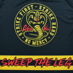 The circular yellow and black Cobra Kai logo featuring a coiled cobra and the words Strike First, Strike Hard, No Mercy above a yellow stripe with the red text Sweep the Leg.