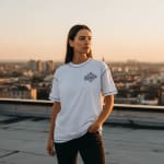 A woman in a white t-shirt with blue stitching and dark pants stands on a rooftop at sunset, looking out over a blurred city skyline.