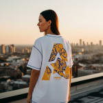 Back view of a woman wearing a white oversized t-shirt with a blue and yellow tiger graphic inside a letter 'A', standing on a rooftop with a city skyline at sunset.