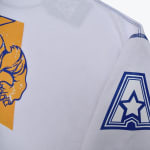 Close-up of a white long-sleeve shirt showing a blue and yellow graphic on the body and a blue star logo inside an A shape on the sleeve.