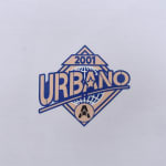 A diamond-shaped logo with the text URBANO in blue and beige, featuring 2001 at the top, a globe in the background, and a circular A star emblem at the bottom, printed on white fabric.