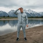 A man in a grey tracksuit stands on a sandy lake shore with his hand raised to his forehead, with a calm lake and snow-capped mountains in the background.