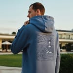 A man wearing a blue hooded sweatshirt with a detailed line-art graphic of a lighthouse on the back, standing outdoors.