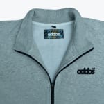 Close-up of a grey zip-up jacket with a ribbed collar, black zipper, and addos embroidered in black on the left chest.
