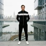 A man standing on a modern balcony wearing a black and white tracksuit and white sneakers, with the New York City skyline in the background.