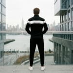 Rear view of a man in a black and white tracksuit standing on a balcony overlooking a city skyline and river.