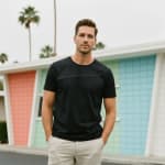A man posing in a black t-shirt and beige trousers in front of a colorful mid-century modern building with palm trees.