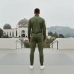 Back view of a man wearing a green tracksuit and white sneakers, standing in front of the Griffith Observatory with the Hollywood Hills in the background.
