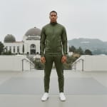 A full-body shot of a man in a matching olive green tracksuit and white sneakers standing on a concrete platform at the Griffith Observatory.