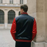 Back view of a man leaning against a stone pillar wearing a black varsity jacket with red sleeves and striped trim.