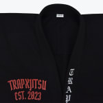 Close-up of a black Jiu-Jitsu gi jacket with red TRAPXJITSU EST. 2023 embroidery on the chest and white vertical TRAP embroidery on the lapel.