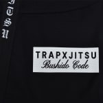 A close-up of a black Jiu-Jitsu Gi featuring a white patch with TRAPXJITSU Bushido Code text and vertical gothic lettering on the labe.