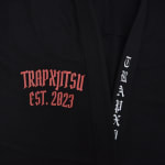 Close-up of a black Jiu-Jitsu gi with red TRAPXJITSU EST. 2023 embroidery on the chest and white vertical lettering on the lapel.