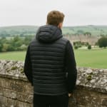 Rear view of a man in a black hooded puffer vest looking out over a green countryside landscape with a stone manor house in the distance.