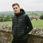 A man in a black hooded puffer vest with PRAGMATIC printed on the chest, standing by a stone wall with a scenic green landscape in the background.