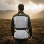 Rear view of a man wearing a dark green and white mesh Hawkvest shooting vest, standing in a grassy field with mountains in the background under a sunset sky.