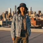 A young woman in a denim jacket, bucket hat, and cargo pants posing on a rooftop with a city skyline in the background during golden hour.
