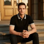 A man in a black polo shirt with grey collar trim sits on stone steps with his hands clasped, looking at the camera during golden hour.