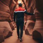 Back view of a man in a black and red tracksuit walking through a narrow orange sandstone slot canyon.