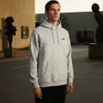 A man wearing a light grey heathered hoodie stands in an outdoor courtyard with modern sculptures in the background under warm golden hour light.