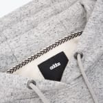 Close-up of a grey heathered hoodie inner neckline, showing a black tag with the brand name addos, a patterned neck tape, and drawstrings.