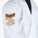 Close-up of the sleeve of a white martial arts gi, featuring a textured weave and an embroidered patch with brown and beige chevrons and the Addos brand name.