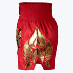 Side view of red satin Muay Thai shorts with a thick elastic waistband and a metallic gold flame design embroidered on the leg against a white background.