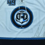 Close-up of a light blue hockey jersey featuring a circular logo with a lighthouse, anchor, crossed hockey sticks, and the letters SCK, with the number 49 on the chest.
