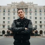 A man in a black and grey varsity jacket with arms crossed, standing in front of the Beverly Wilshire hotel building.