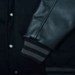 Close-up of a black varsity jacket showing the textured leather sleeve, striped ribbed cuff, wool body, and custom snap buttons.
