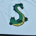 A stylized green sea serpent shaped like the letter S with a yellow underbelly and dark green fins, printed on a light blue fabric background.