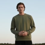 A man wearing a solid olive green crewneck sweatshirt stands outdoors with his hands clasped, looking at the camera against a clear sky.
