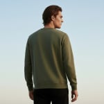 Back view of a man wearing a long-sleeved olive green sweatshirt and black pants against a clear blue sky.