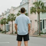 Back view of a man in a light blue short-sleeve shirt and black shorts standing on a street with pastel-colored historic houses and palm trees in the background.