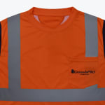 Close-up of an orange high-visibility safety shirt with reflective silver stripes and a pocket featuring the GroundsPRO logo.