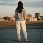 Back view of a woman in a light blue t-shirt and white trousers standing on a rooftop looking at a city skyline at sunset.