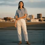 A woman stands on a rooftop wearing a light blue polo shirt and white trousers with a city skyline behind her.