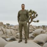 A man in olive green long-sleeve shirt and cargo pants stands on a large boulder in a desert filled with Joshua trees and rock formations under a cloudy sky.