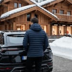 Rear view of a man in a navy blue quilted puffer jacket standing behind a black luxury SUV in a snowy alpine village with wooden chalets.