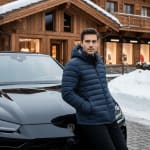 A man in a blue down jacket leaning on a black luxury car in front of a wooden chalet in a snowy setting.