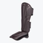 A side-angle view of a dark brown leather martial arts shin guard with attached foot protector, featuring white stitching, on a white background.