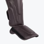A dark brown leather shin and instep guard with white stitching, displayed against a plain white background.