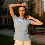 A woman posing in a light blue polo shirt and white pants outdoors during sunset, with a blurred building in the background.