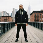 A man stands in the center of a wooden elevated walkway wearing a black hoodie with neon yellow details and black sweatpants, with the New York City skyline in the background.