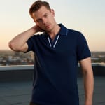 A young man with styled brown hair wearing a navy blue polo shirt stands on a rooftop at sunset, posing with one hand behind his neck.