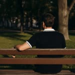 Rear view of a man wearing a black polo shirt sitting on a wooden bench in a sunny park.