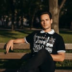 A man in a black graphic polo shirt sitting on a wooden bench in a park during sunset.