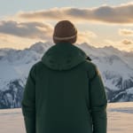 Back view of a person in a green winter coat and brown beanie looking out at a vast range of snow-covered mountains during a golden sunset.